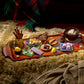 A cozy holiday-themed food and drink spread arranged on a wooden tray over a faux-fur surface, with a plaid blanket and knit sweater in the background. The tray holds a mug of hot chocolate, pastries, cookies, chocolates, dice, and small decorative items.