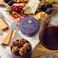 A round purple tin of hand-rolled chocolate truffles sits on a wooden charcuterie board surrounded by cheese, blackberries, pomegranate, rosemary, and a glass of red wine. A smaller purple container in front holds several truffles, with wooden-handled knives nearby.