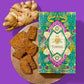 A green and purple gable box sits beside several pieces of Gingerbread Toffee on a wooden tray beside a large ginger root on a bright purple background.