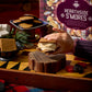 Close-up of a hand pressing a graham cracker onto a marshmallow with the Hearthside S'mores box in the background.