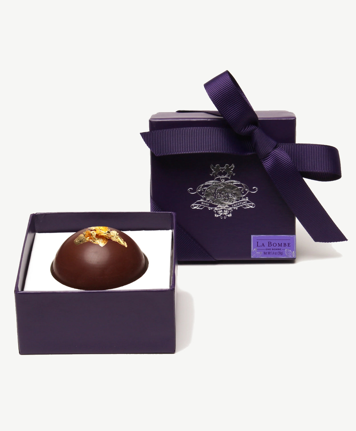 A large hazelnut Reishi mushroom chocolate bombalina truffle in an opened purple candy box standing upright tied with a purple ribbon bow on a white background.