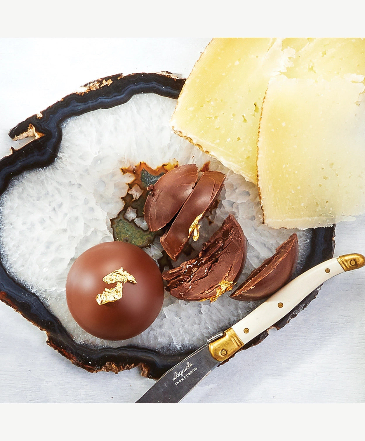 Two Vosges bombalina la bombe chocolate Reishi hazelnut ganache truffles topped in gold leaf sit atop a crystal geode beside a serving knife and two slices of cheese on a white background.