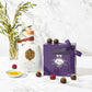 A bottle of Kyoord olive oil tied with a leather strap sits beside a purple candy box and a dish of oil surrounded by brightly decorated chocolate truffles on a marble background.
