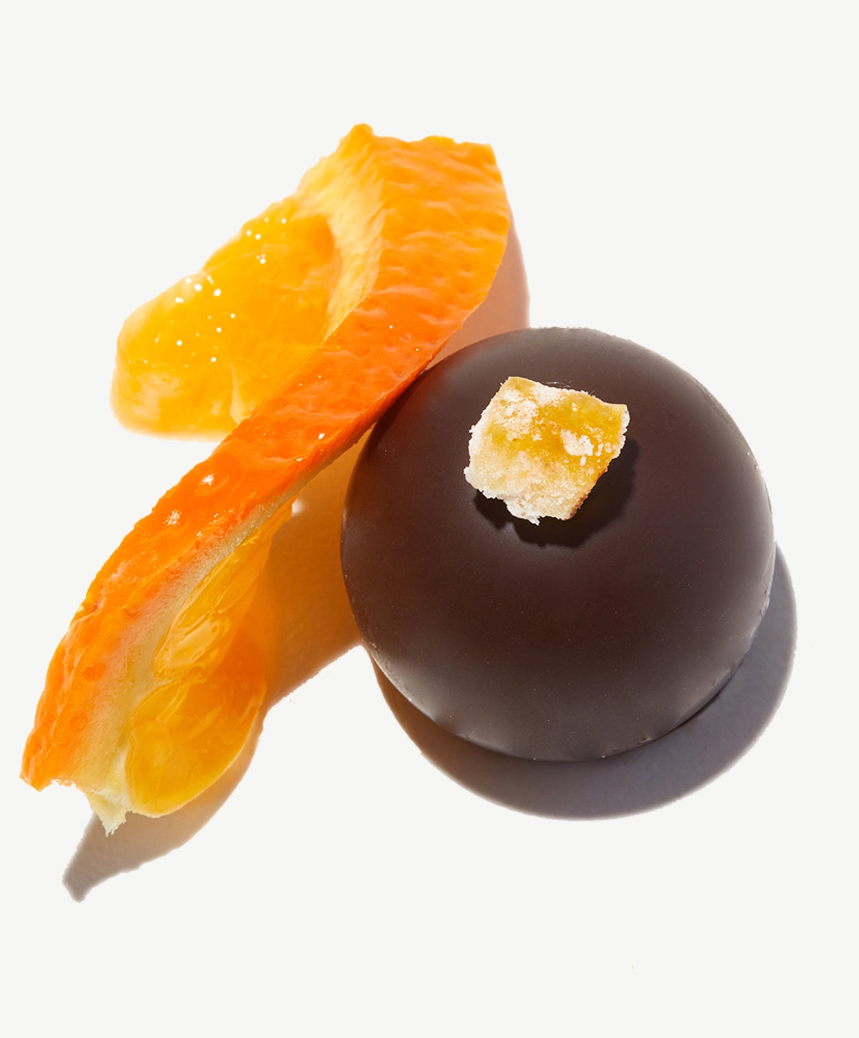 Close-up of a Vosges Sicilian Blood Orange vegan truffle next to a slice of fresh orange topped with a candied orange peel on a white background.