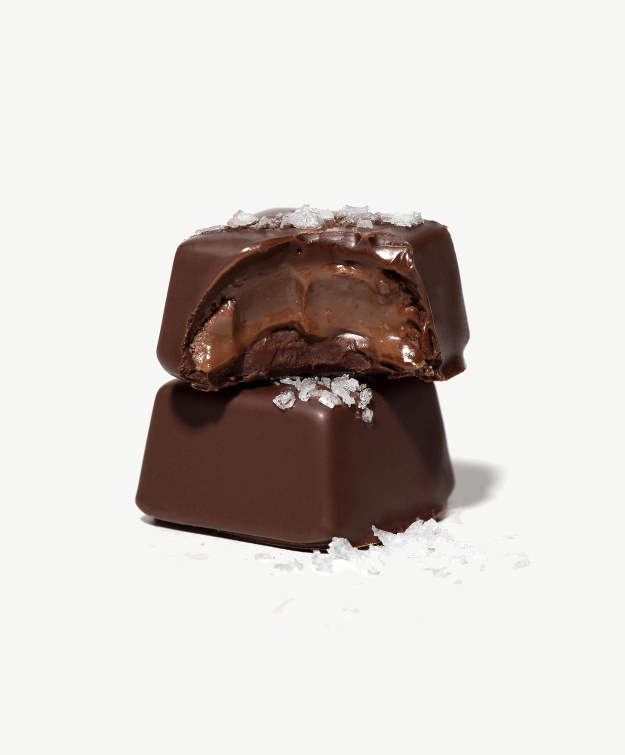 Close-up of two Vosges Peanut Butter Bonbons, one with a bite taken displaying a creamy peanut butter chocolate center topped with sea salt on a white background.