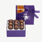 Open purple candy box tied embossed in silver tied with a purple ribbon bow sits upright displaying nine Vosges milk chocolate praline truffles topped with gold leaf, candied violet and coco nibs on a white background.