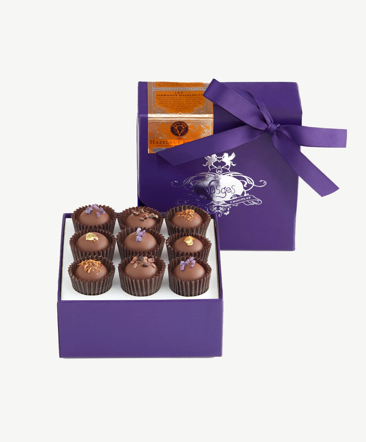 Open purple candy box tied embossed in silver tied with a purple ribbon bow sits upright displaying nine Vosges milk chocolate praline truffles topped with gold leaf, candied violet and coco nibs on a white background.