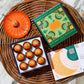 A box of Vosges Pumpkin truffles sits open on a wicker tray beside a bright orange desert plate containing a vosges truffle topped with a pumpkin seed on a grey marble background.