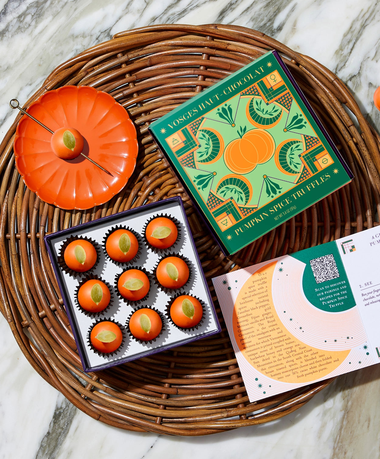 A box of Vosges Pumpkin truffles sits open on a wicker tray beside a bright orange desert plate containing a vosges truffle topped with a pumpkin seed on a grey marble background.