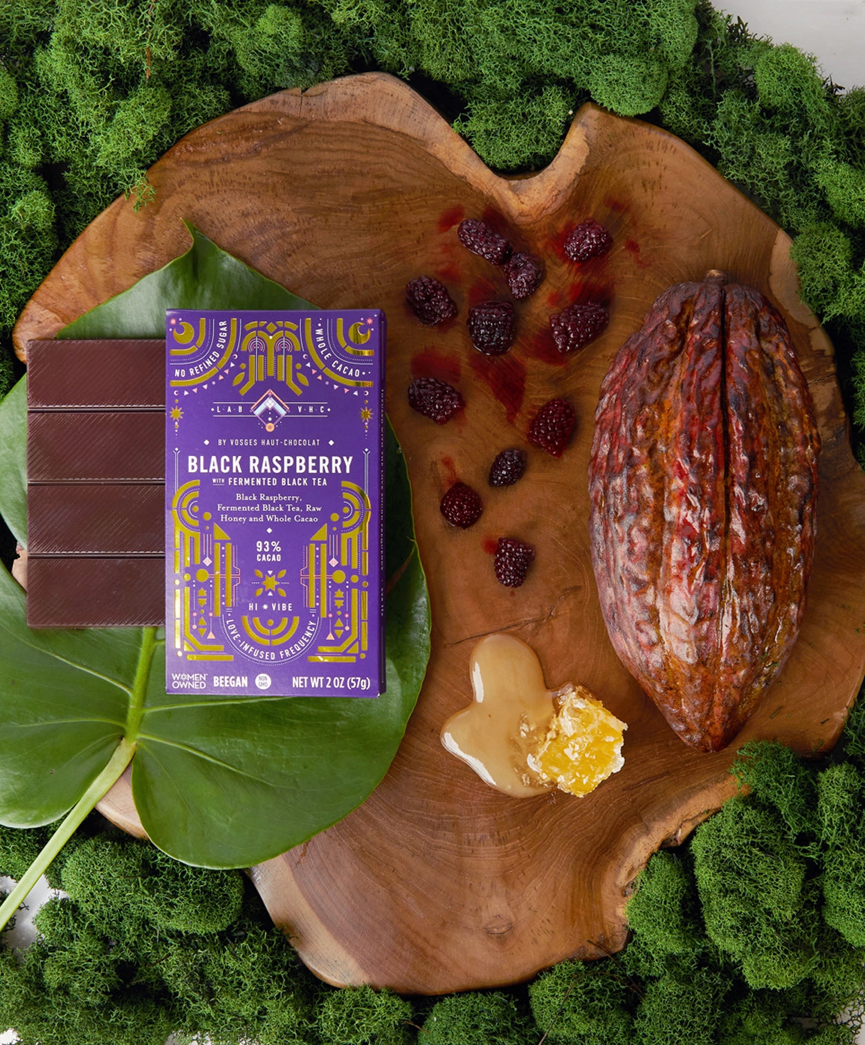 Vosges Black Raspberry Bar on a large green leaf beside a cacao pod surrounded by green moss, honey comb and fresh raspberries.