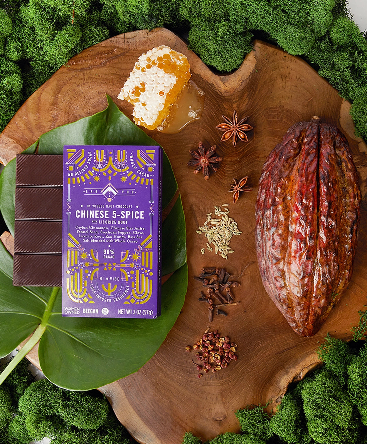 Vosges Chinese 5-Spice Bar on a large green leaf beside a cacao pod surrounded by green moss, honey comb and spices.