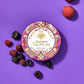 A round Vosges chocolate box decorated in bright red, pink and gold foil sits beside several fresh raspberries and Vosges Raspberry truffles, on a light purple background.