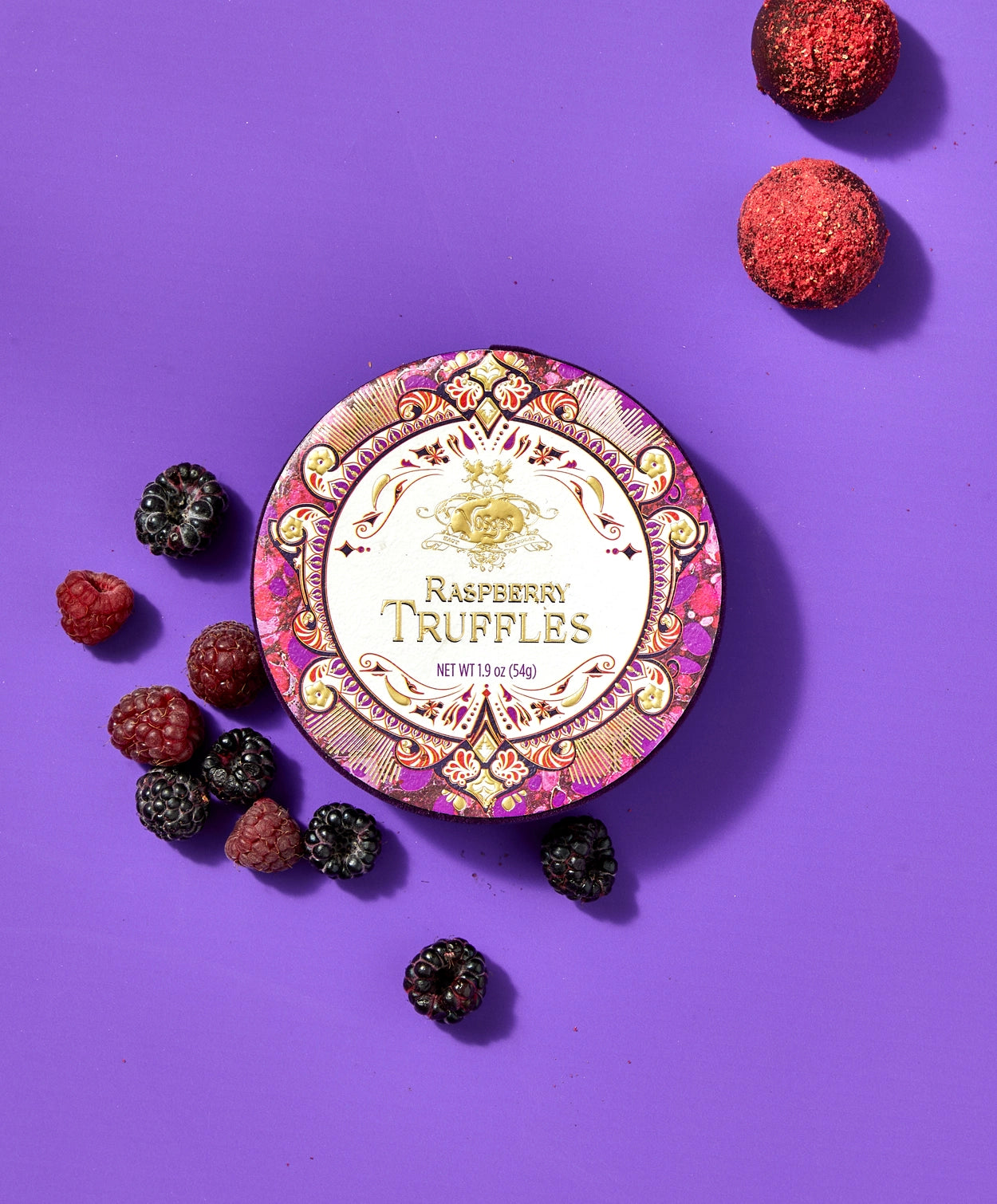 A round Vosges chocolate box decorated in bright red, pink and gold foil sits beside several fresh raspberries and Vosges Raspberry truffles, on a light purple background.