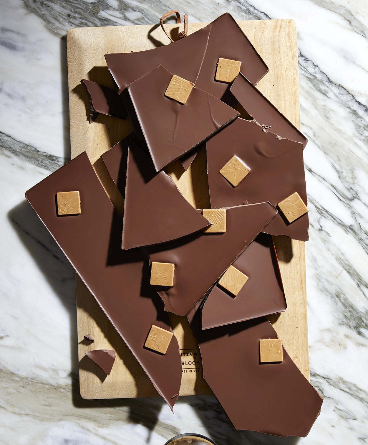 Several broken pieces of Vosges Soiree Smash on a wooden cutting board sitting on a marble countertop.  