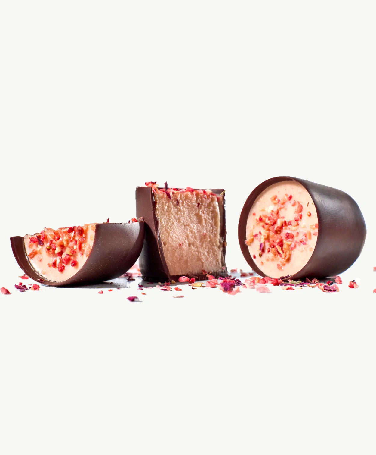 Two Vosges Strawberry Mascarpone Desert cups, one cut in half revealing pink sweet cheese in a chocolate coating topped with dragon fruit on a bed of dried rose petals on a grey background.