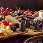 A wooden charcuterie board filled with cheeses, crackers, grapes, berries, and several chocolate-covered cone-shaped snacks on round crackers, photographed in a dark rustic setting.