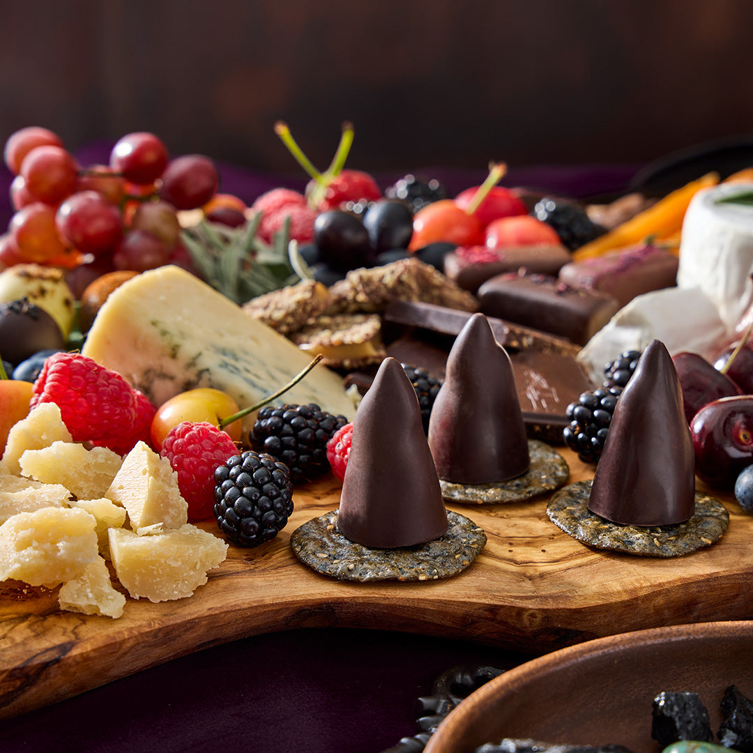 A wooden charcuterie board filled with cheeses, crackers, grapes, berries, and several chocolate-covered cone-shaped snacks on round crackers, photographed in a dark rustic setting.