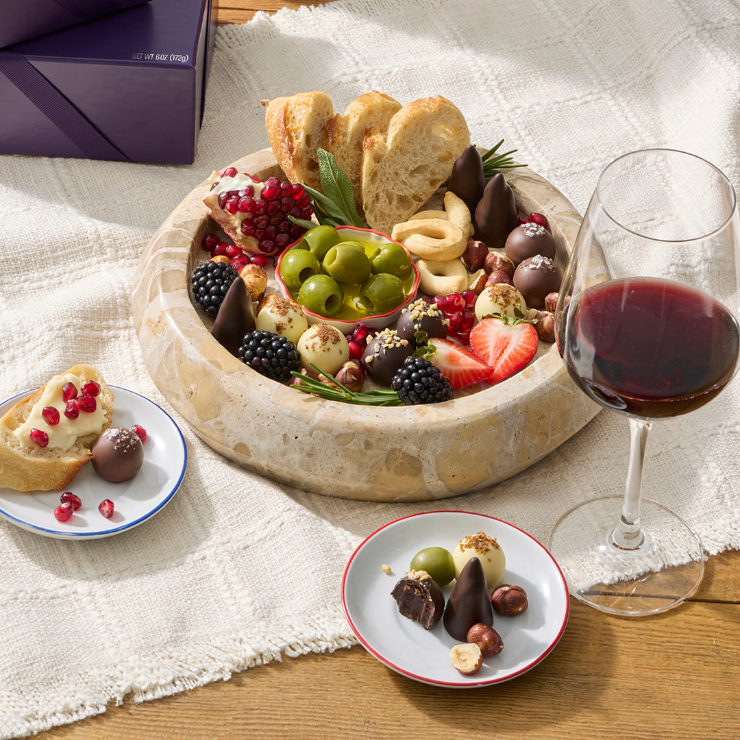 A gourmet charcuterie board or cheese board arrangement in a round stone dish, styled with sliced baguette, pomegranate, green olives, strawberries, blackberries, nuts, chocolates, and a glass of red wine on a cream tablecloth.