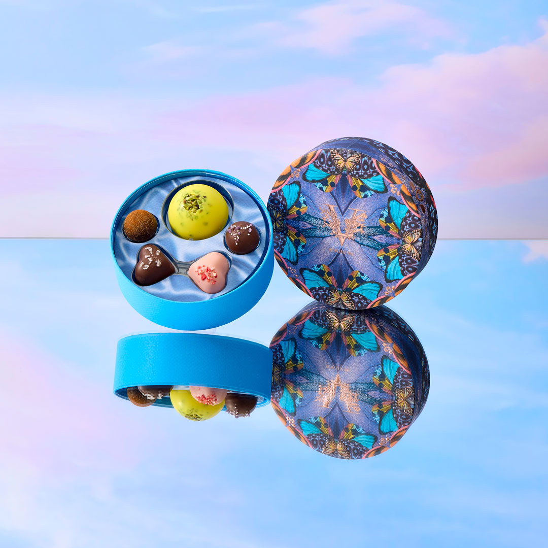 Decorative chocolate tin with colorful butterfly design on a reflective surface with a pastel sky background.