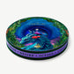 A circular Vosges chocolate collection box trimmed in purple velvet, decorated with a blue dragon and pink egret in a green forest in front a dark nighttime sky, on a light grey background. 