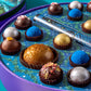 Close up of several Vosges Chocolate truffles adorned in brightly colored powders and ingredients, in an opened candy box decorated as a night sky  sits open on a light blue background.