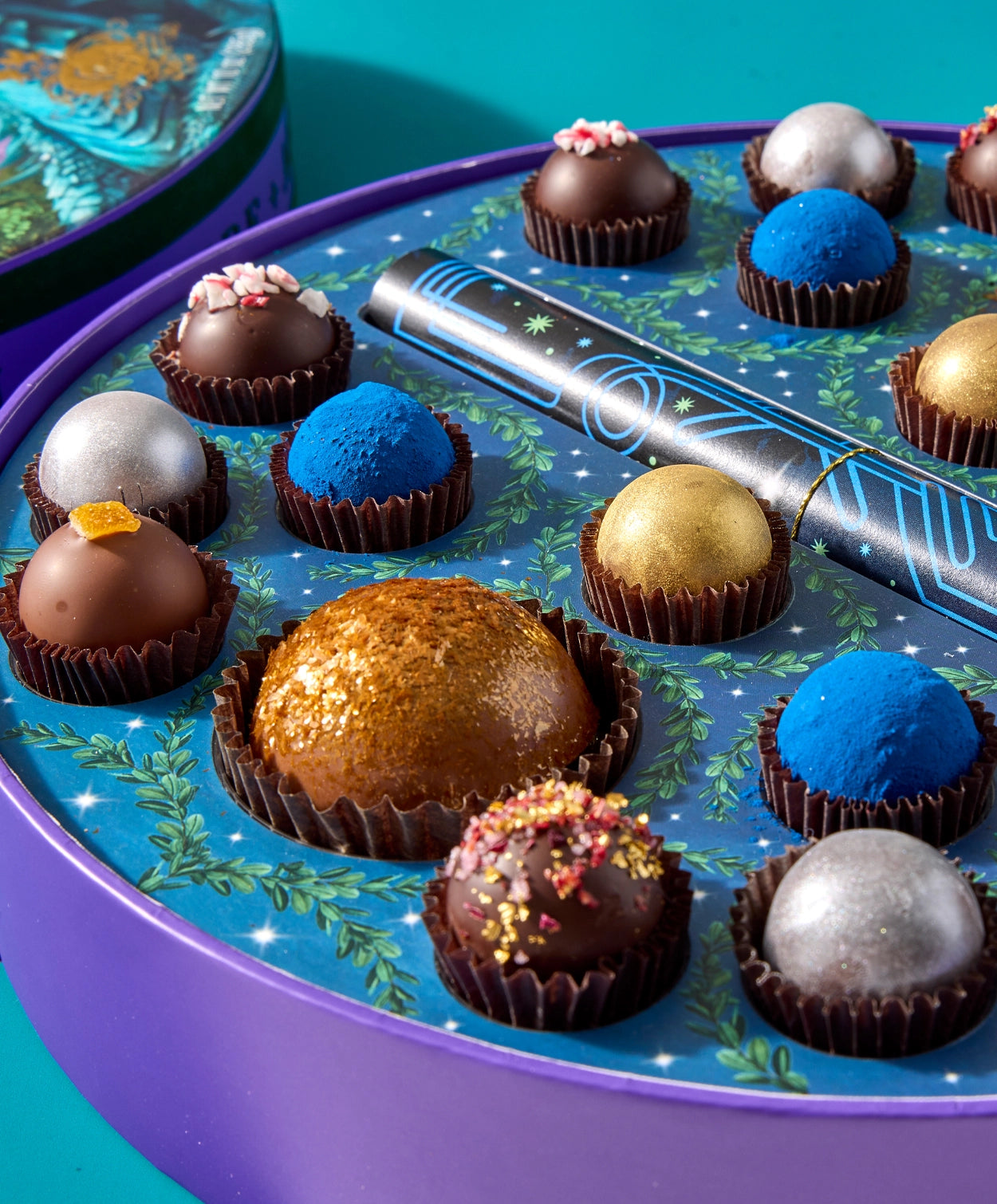 Close up of several Vosges Chocolate truffles adorned in brightly colored powders and ingredients, in an opened candy box decorated as a night sky  sits open on a light blue background.