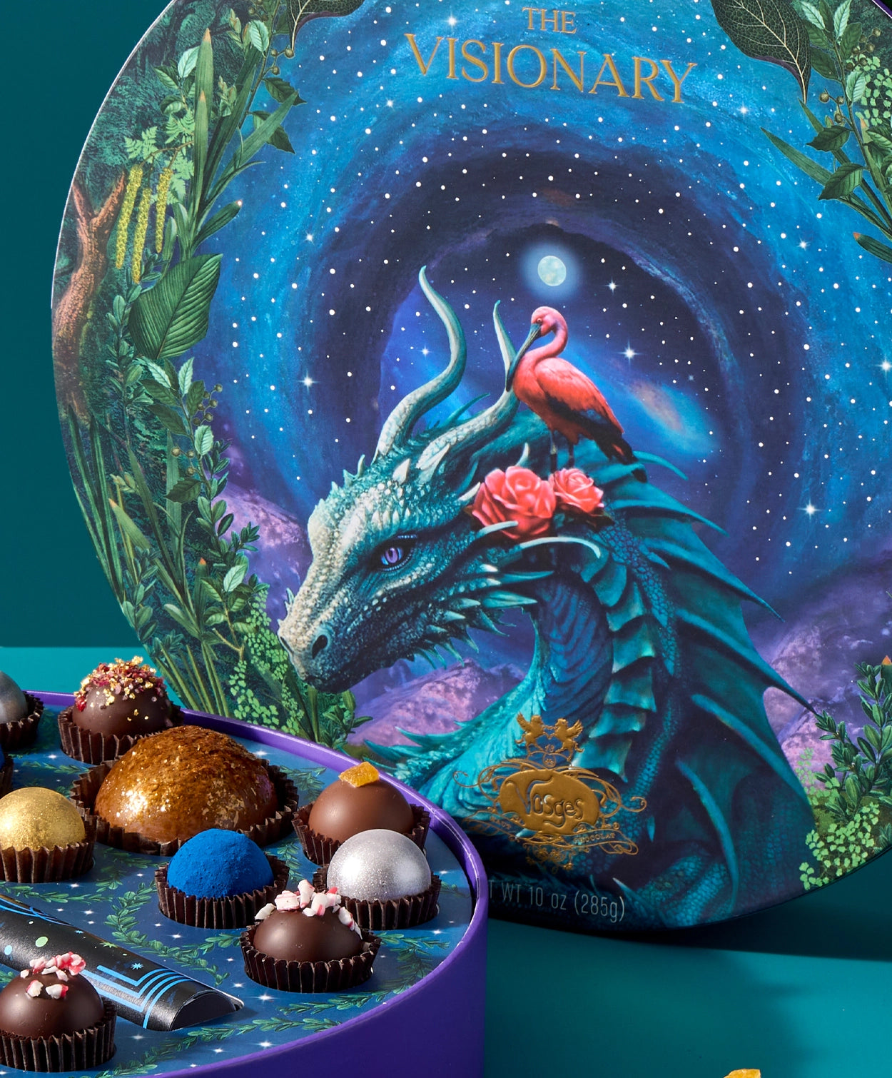A closeup view of the Vosges Visionary collection artwork, ,a large blue dragon and pink egret sitting in a lush leafy forest infront a swirling night sky full of stars.