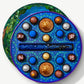 A round Vosges chocolate box sits open atop it's densely decorated lid, revealing twenty brightly colored chocolate truffles adorned in blue algae and crushed peppermints, on a light grey background.