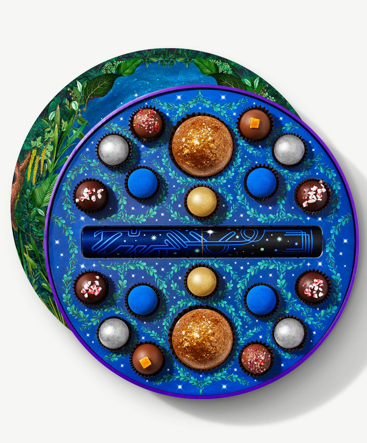 A round Vosges chocolate box sits open atop it's densely decorated lid, revealing twenty brightly colored chocolate truffles adorned in blue algae and crushed peppermints, on a light grey background.