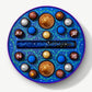 A round Vosges chocolate box sits open, revealing twenty brightly colored chocolate truffles adorned in blue algae and crushed peppermints, on a light grey background.