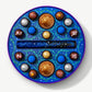 A round Vosges chocolate box sits open, revealing twenty brightly colored chocolate truffles adorned in blue algae and crushed peppermints, on a light grey background.