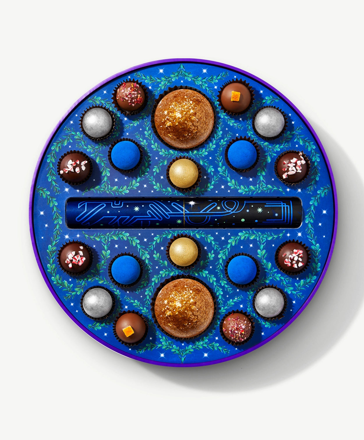 A round Vosges chocolate box sits open, revealing twenty brightly colored chocolate truffles adorned in blue algae and crushed peppermints, on a light grey background.