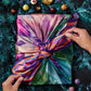 a caucasian woman's hands firmly tie a colorful silk scarf around a rectangular chocolate box on a green marble background/