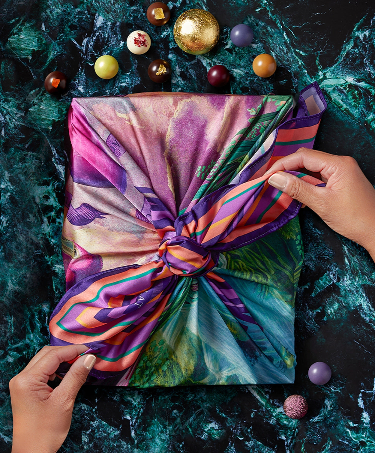 a caucasian woman's hands firmly tie a colorful silk scarf around a rectangular chocolate box on a green marble background/