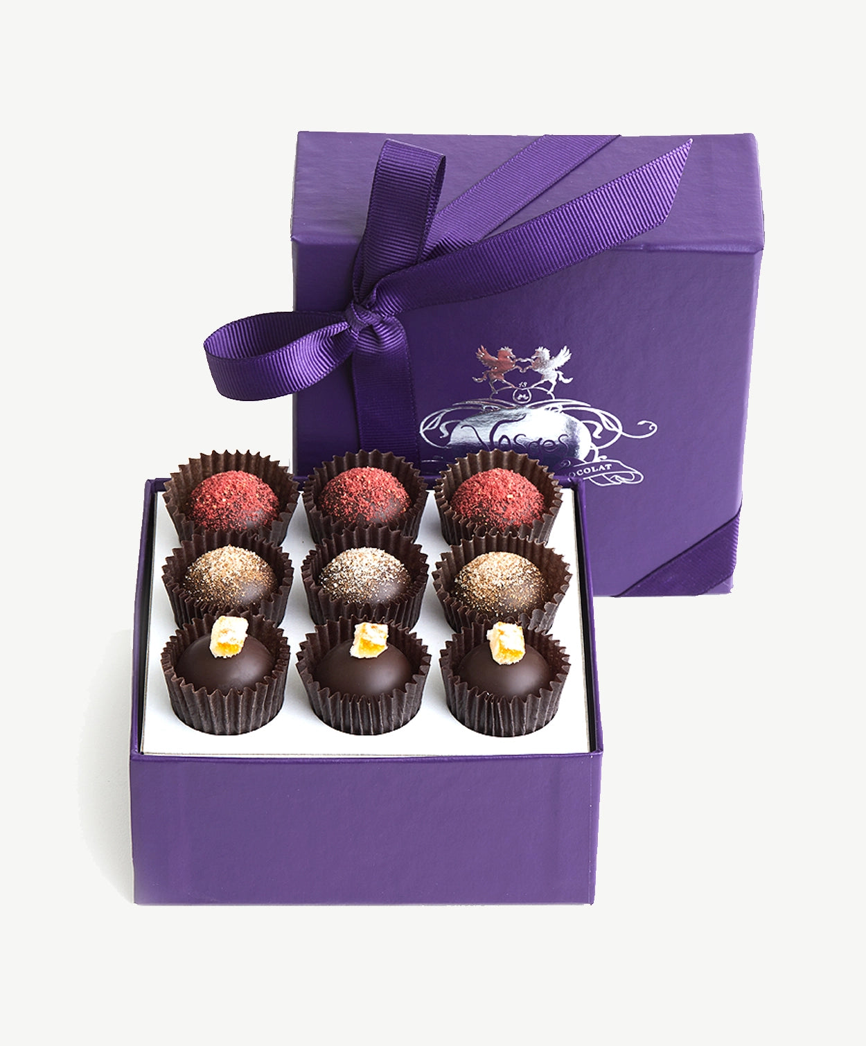 A small purple candy box tied with a purple ribbon bow sit's upright displaying three rows of vegan chocolate truffles adorned in colorful toppings on a white background.