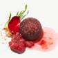 Close-up view of a Vosges vegan chocolate truffle topped with dried raspberries beside a fresh raspberry and flower petals on a white background.