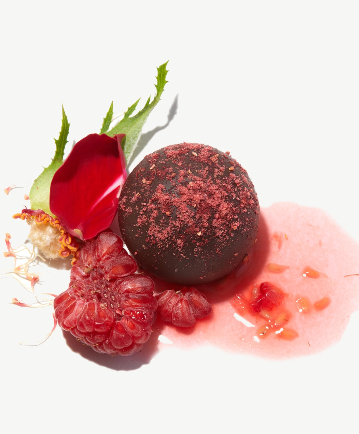 Close-up view of a Vosges vegan chocolate truffle topped with dried raspberries beside a fresh raspberry and flower petals on a white background.