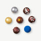 Seven Vosges truffles, decorated with metallic powders, chopped candy cane and bright blue ingredients sit in a hexagon on a light grey background.
