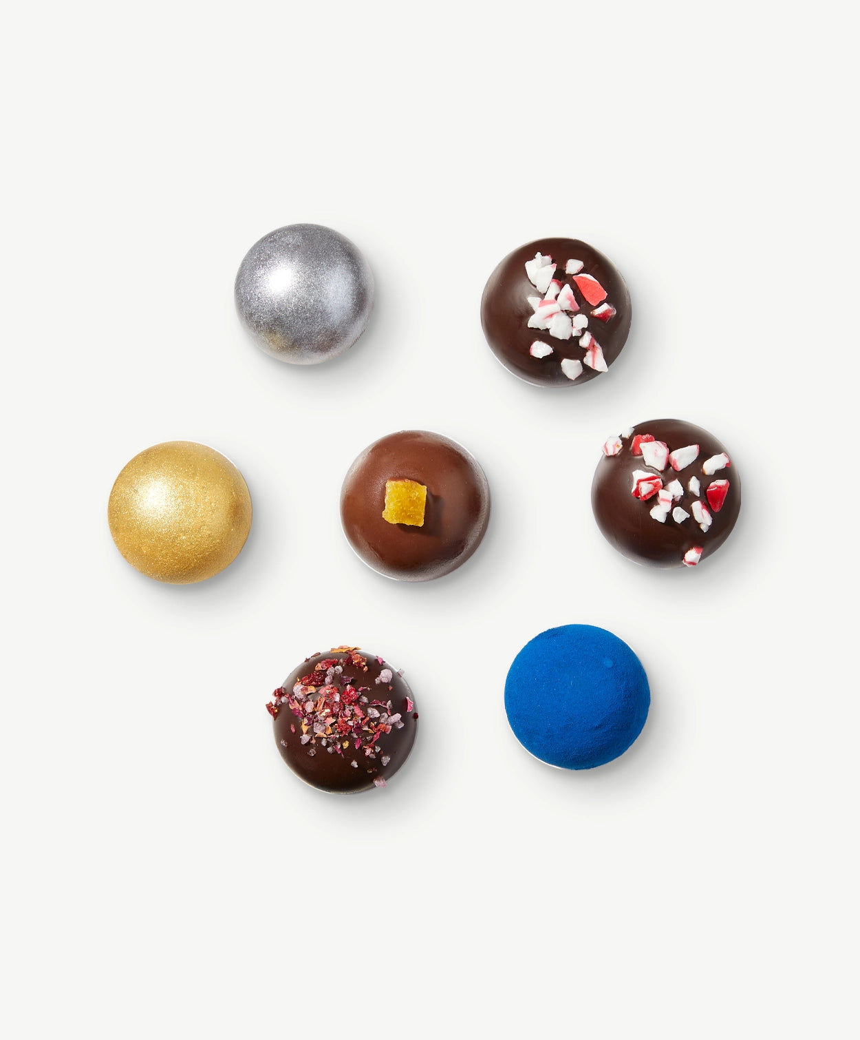 Seven Vosges truffles, decorated with metallic powders, chopped candy cane and bright blue ingredients sit in a hexagon on a light grey background.