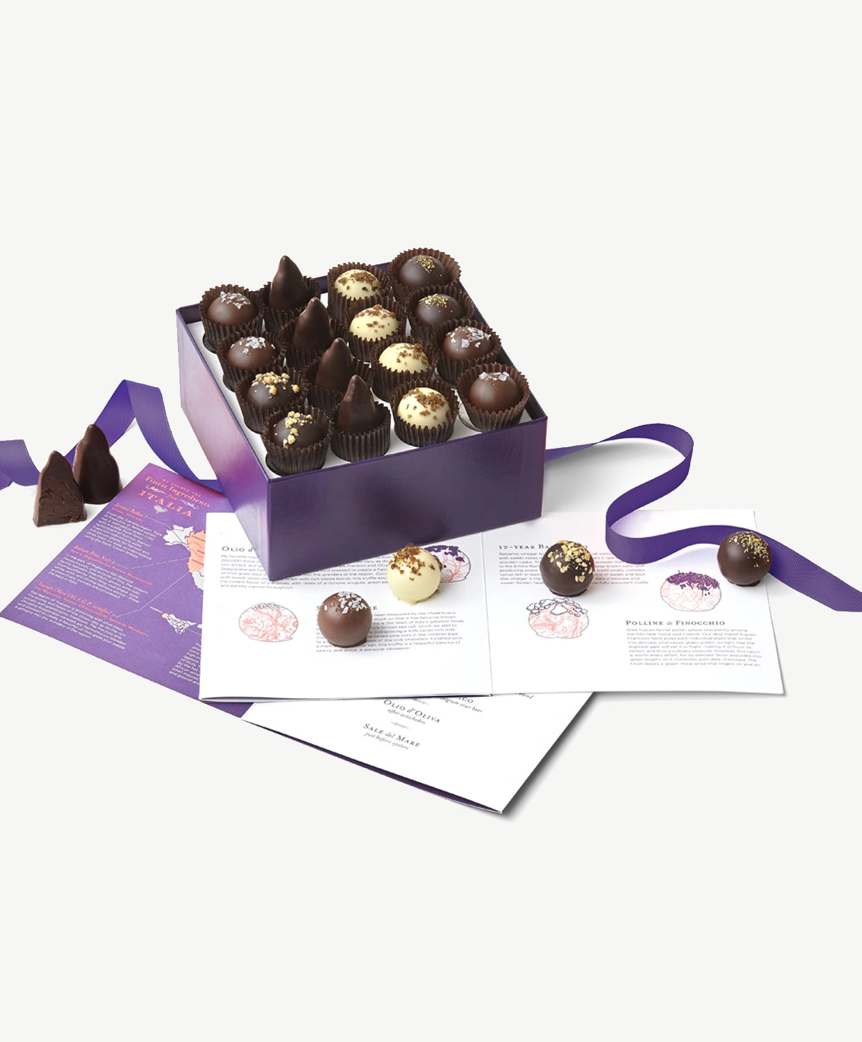 Purple candy box sits open displaying a Voges Italian Truffle collection with sixteen dark, milk and white chocolate bonbons beside several truffles and included tasting guide book on a white background.  