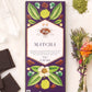 Vosges Matcha Green Tea Spirulina chocolate bar beside a bundle of dried flowers, green matcha powder, and several crystals on a white background.