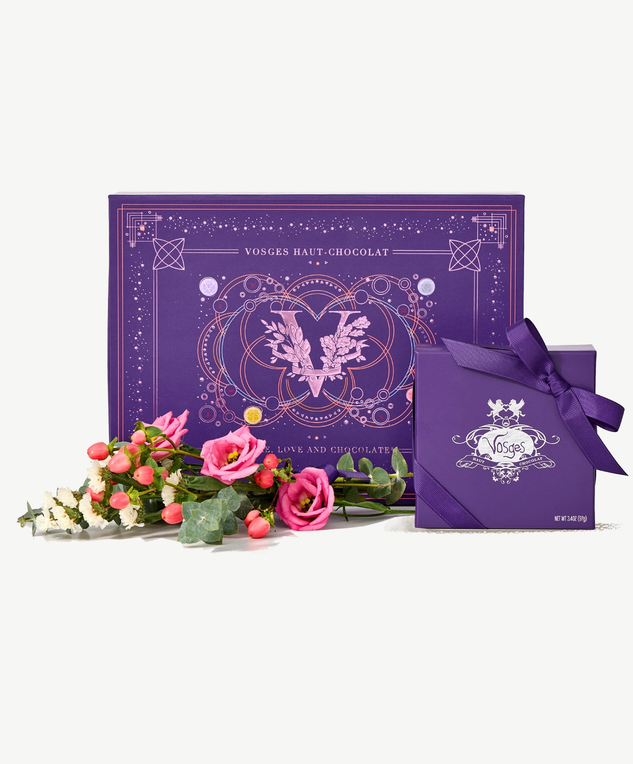 A bouquet of pink flowers sit beside two ornately decorate purple Vosges chocolate boxes on a light grey background.