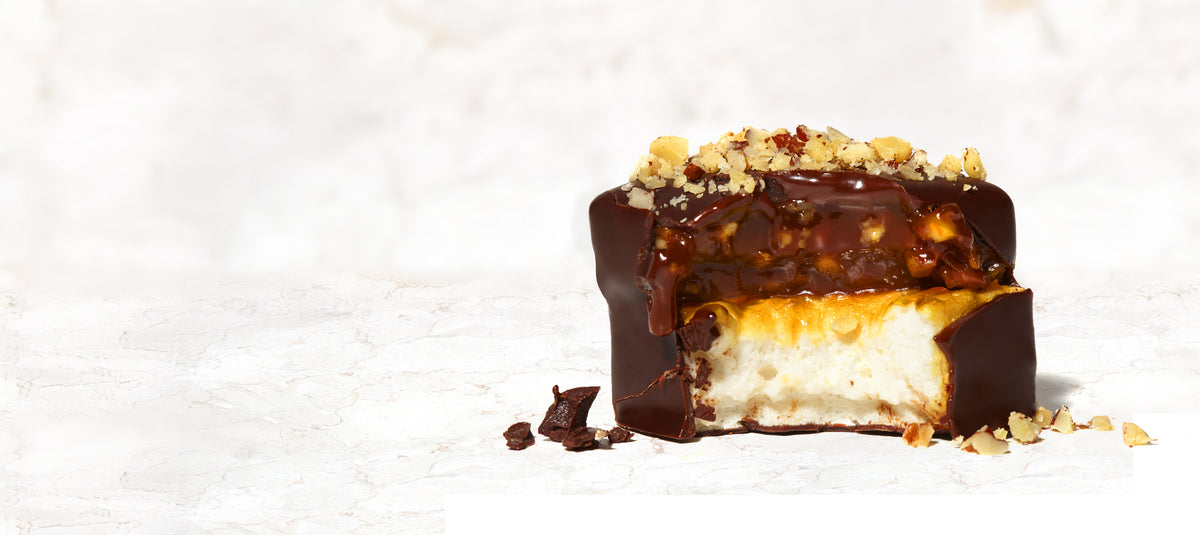 A caramel marshmallow sprinkled with walnut pieces and a bite taken from it on a marble background.