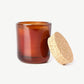A white Linnea’s Lights scented candle in a brown glass jar with a natural cork wood lid leaning against, on a white background.