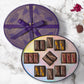 Vosges Exotic Caramel collection sits open displaying eighteen chocolate enrobed caramels beside four piles of colorful nuts and toppings on a marble countertop.