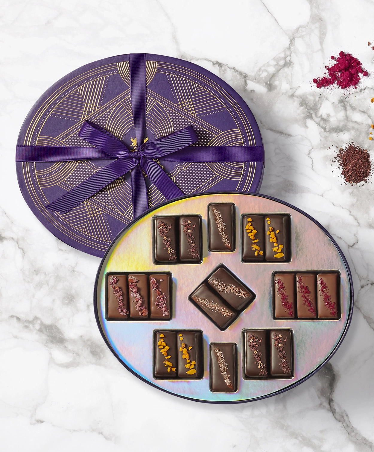 Vosges Exotic Caramel collection sits open displaying eighteen chocolate enrobed caramels beside four piles of colorful nuts and toppings on a marble countertop.