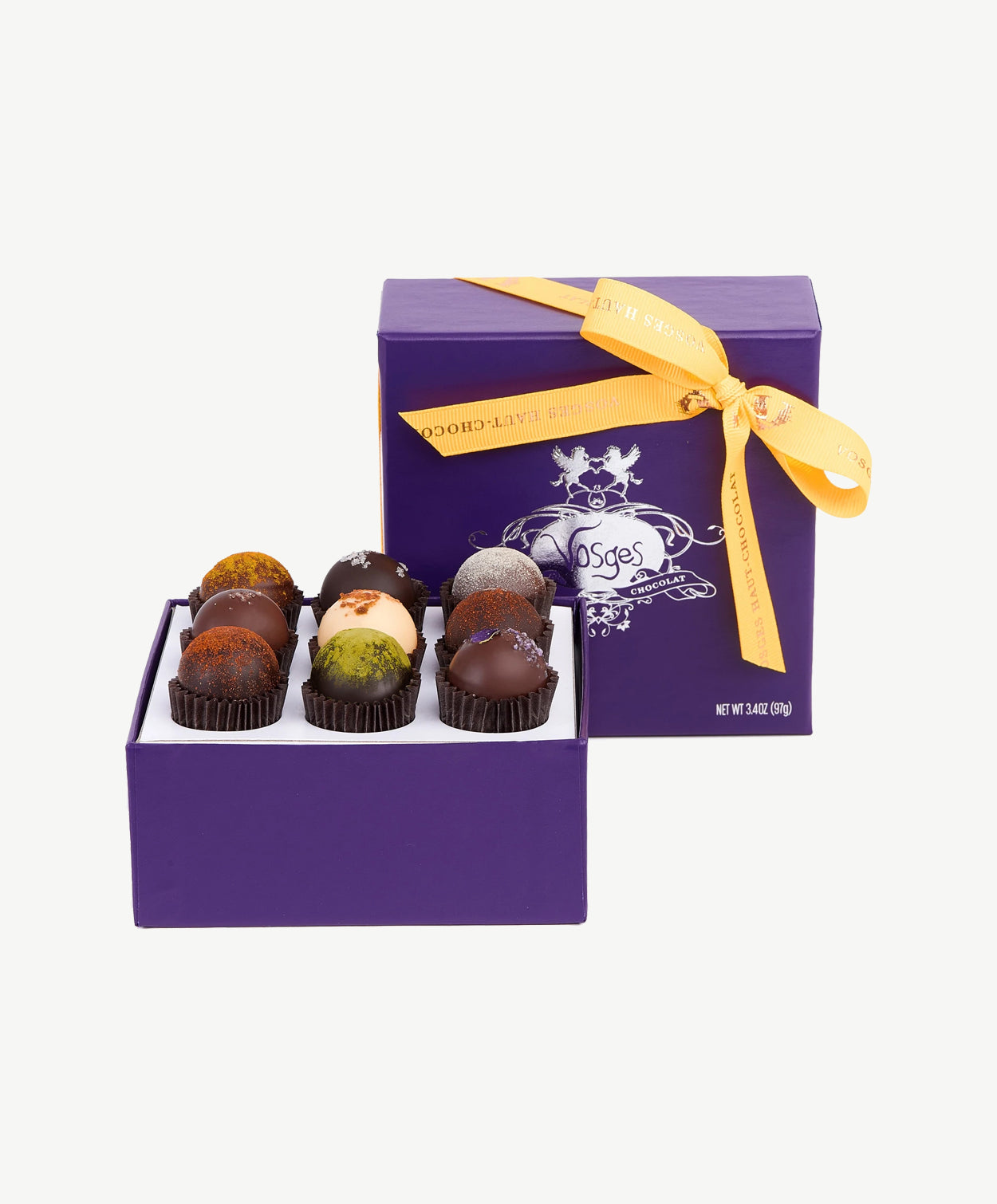 A purple box of Vosges Mindful Truffles sit's open, the lid upright emblazoned with a silver embossed logo tied with a pink bow on a white background. 
