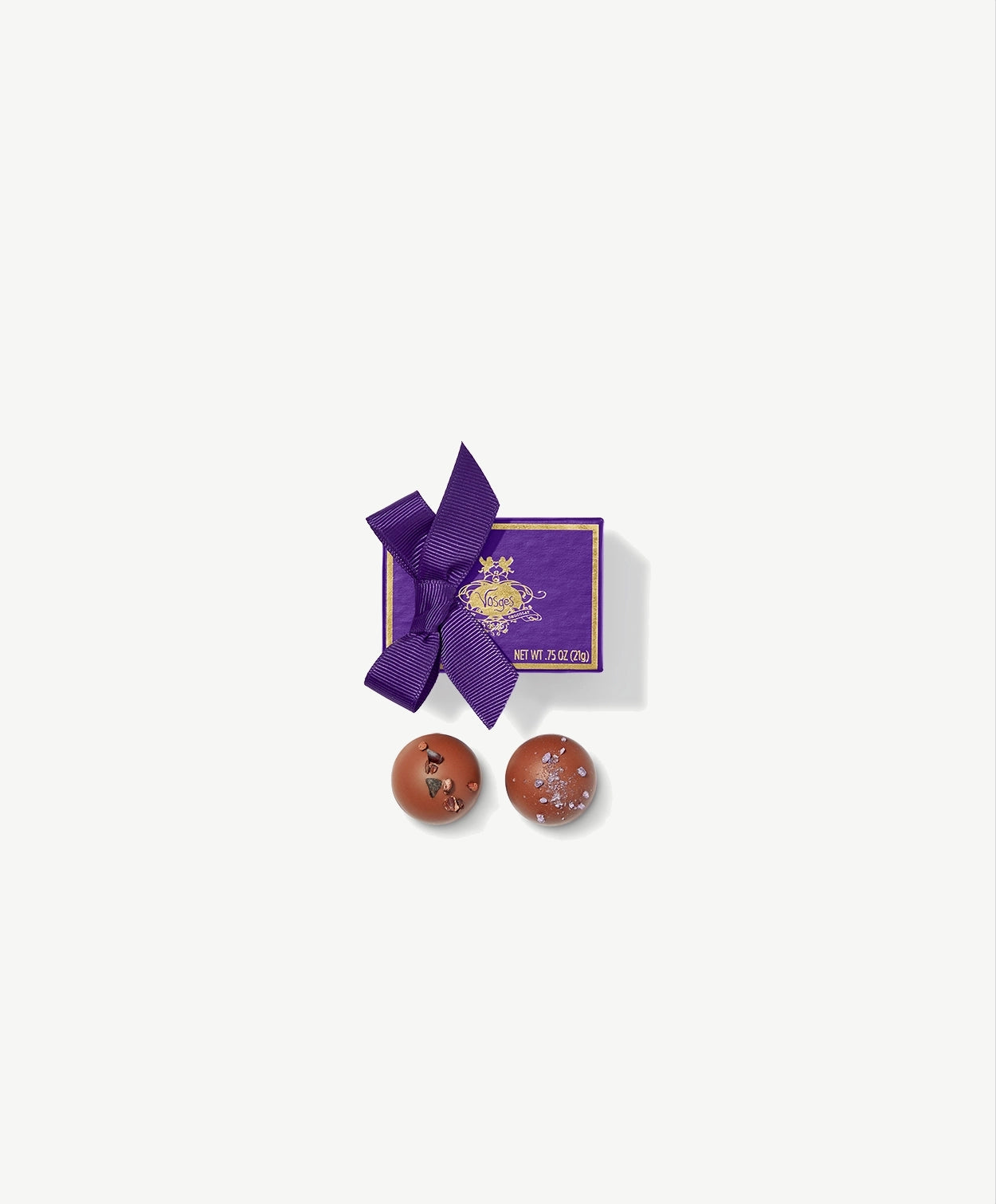 Two Vosges Praline Truffles adorned with Piemonte hazelnuts and candied violet sit side by side in-front a small purple candy box tied with a purple ribbon bow on a grey background.