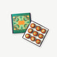 A green and gold chocolate box sits open revealing 9 bright orange pumpkin spice truffles adorned with toasted pumpkin seeds on a light grey backround.