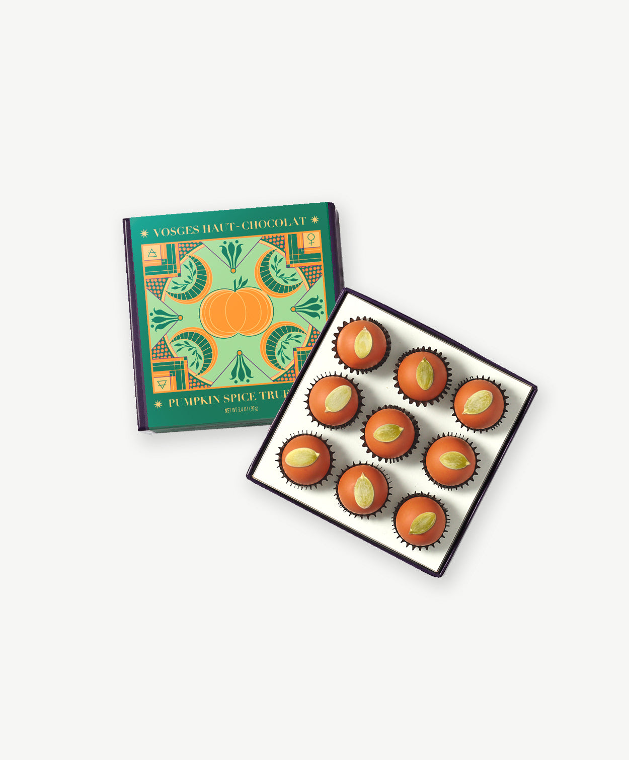 A green and gold chocolate box sits open revealing 9 bright orange pumpkin spice truffles adorned with toasted pumpkin seeds on a light grey backround.
